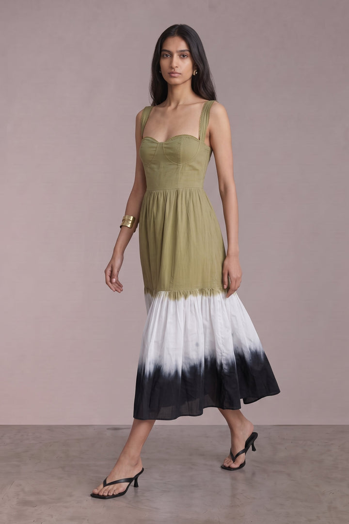A woman modeling a sleeveless midi dress with a structured olive green bodice and a tiered skirt featuring a white-to-black ombre gradient, posed against a solid mauve studio backdrop.