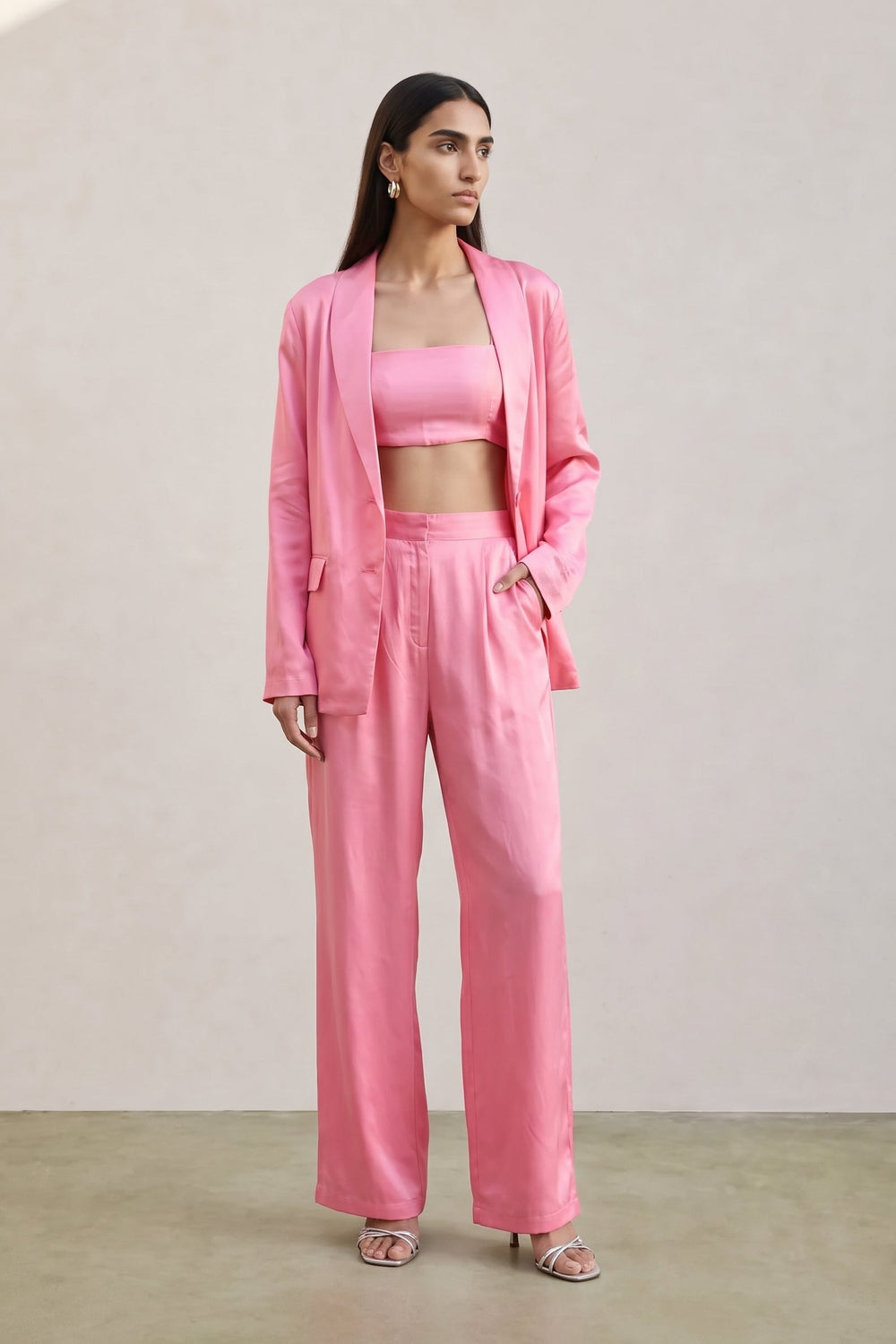 A woman modeling a coordinated pink outfit consisting of a satin-like blazer, matching crop top, and wide-leg trousers, standing against a plain beige wall.