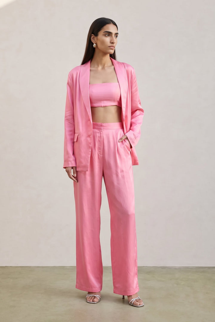 A woman modeling a coordinated pink outfit consisting of a satin-like blazer, matching crop top, and wide-leg trousers, standing against a plain beige wall.
