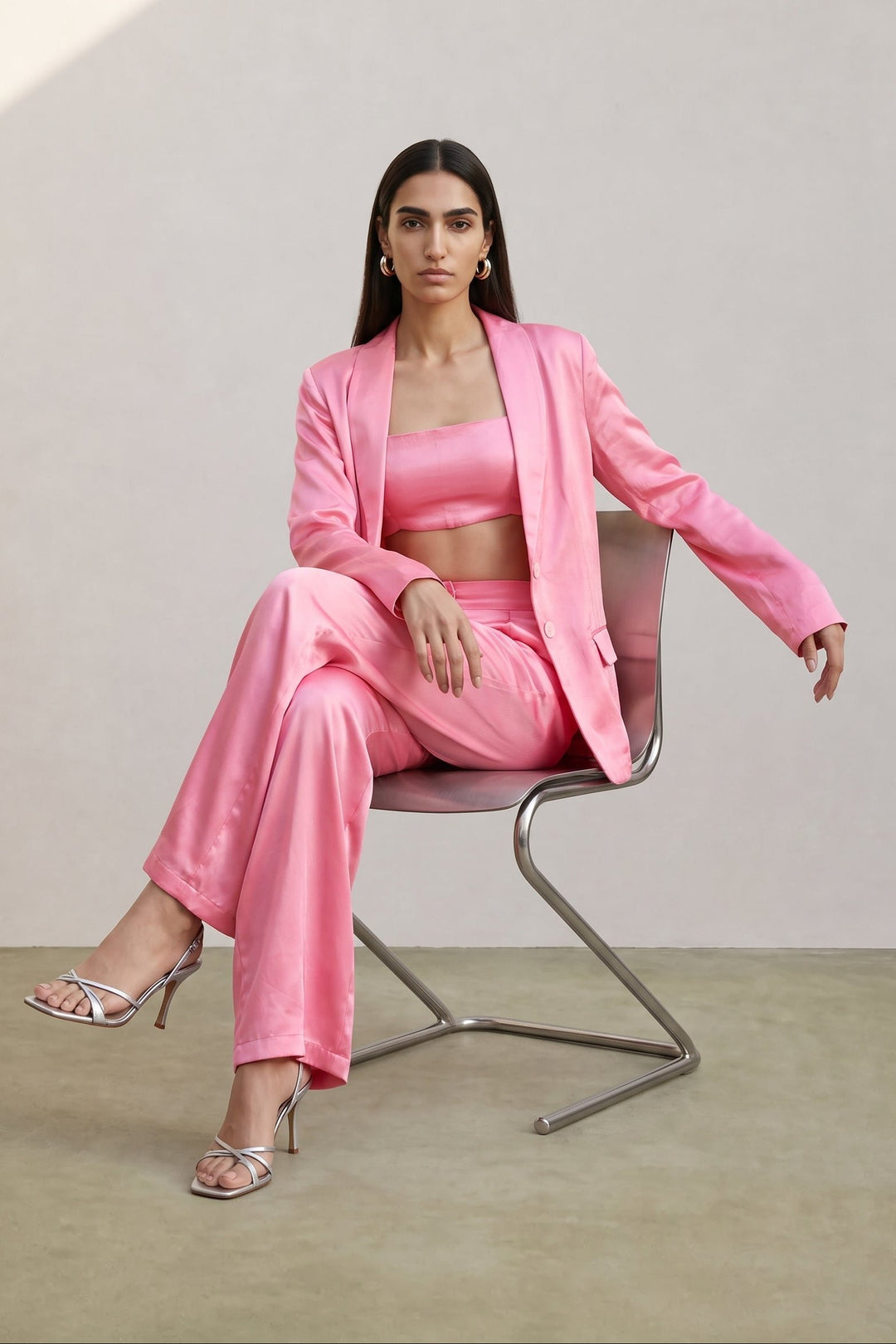 A woman models a matching pink satin three-piece suit consisting of a blazer, crop top, and trousers, seated on a modern metal chair against a neutral beige studio background.
