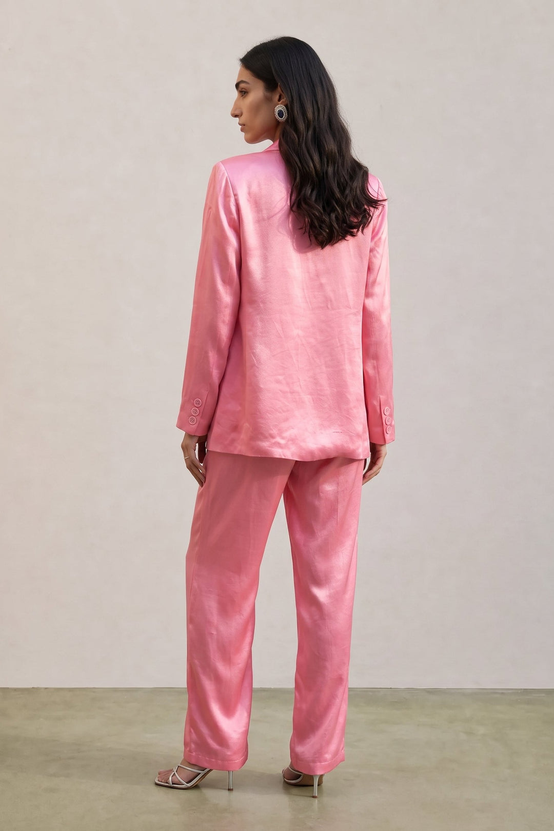 A woman wearing a shiny pink two-piece suit with a blazer and matching trousers, shown from the back against a plain light beige studio wall.