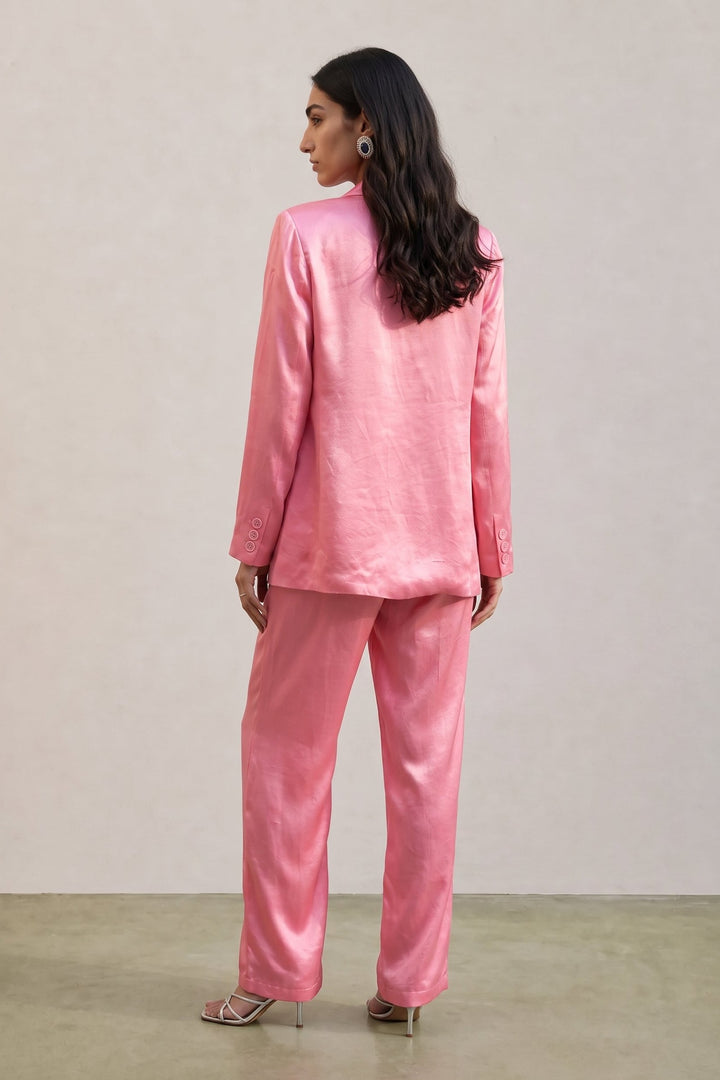 A woman wearing a shiny pink two-piece suit with a blazer and matching trousers, shown from the back against a plain light beige studio wall.
