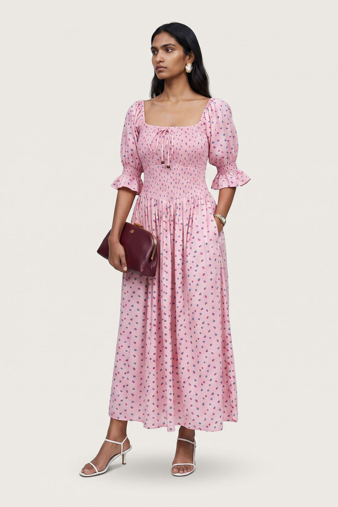 A woman modeling a pink midi dress with a small red and blue floral print, featuring puff sleeves and a square neckline. She is holding a dark burgundy clutch and wearing white strappy high-heeled sandals against a light gray studio background.