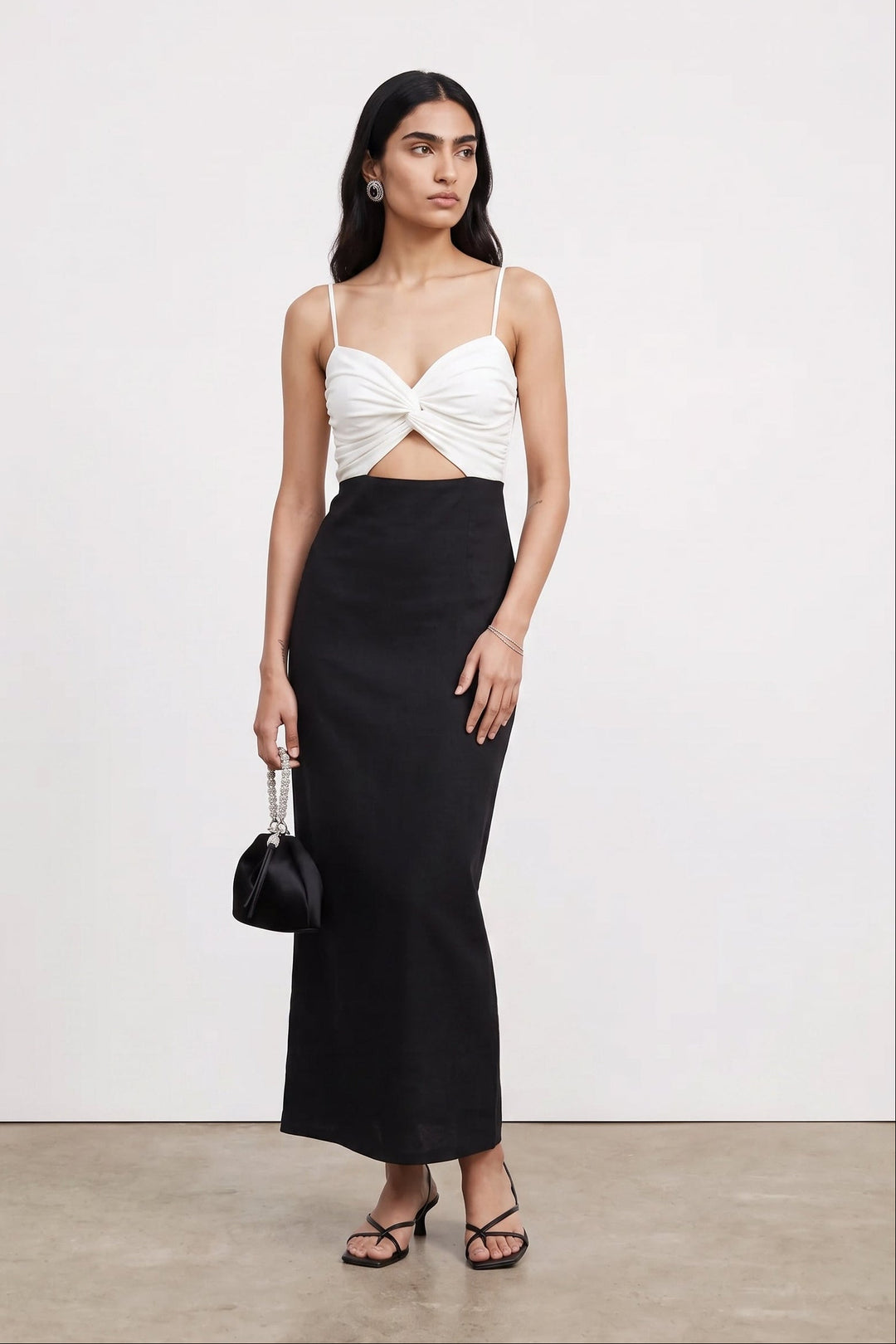 A model stands in a studio wearing a white twisted-front spaghetti-strap crop top and a long black skirt, holding a small black handbag.