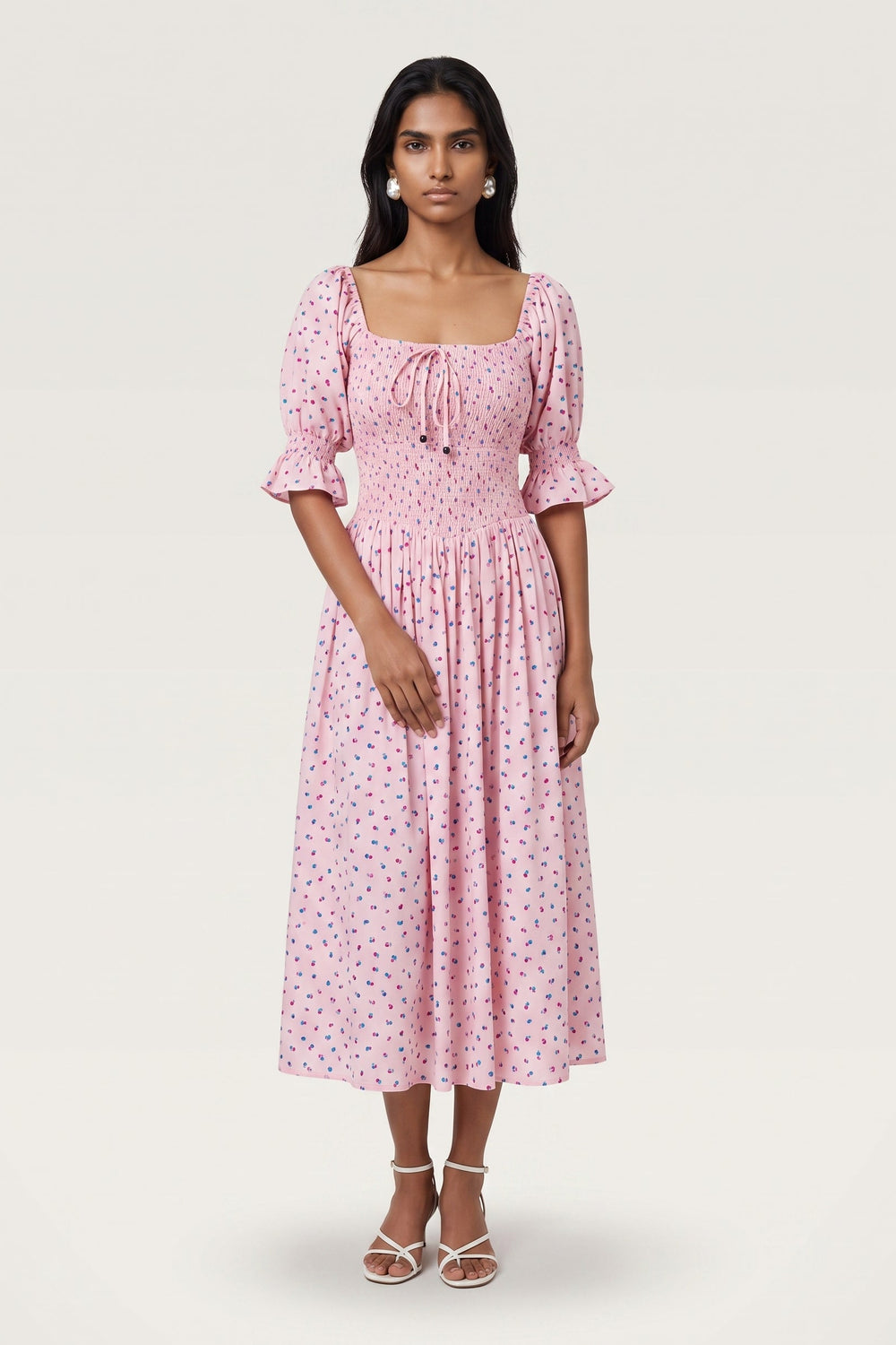A woman modeling a pink midi dress with a small multicolor floral print, featuring puff sleeves, a square neckline with a tie, and a gathered skirt, set against a plain light gray studio background.