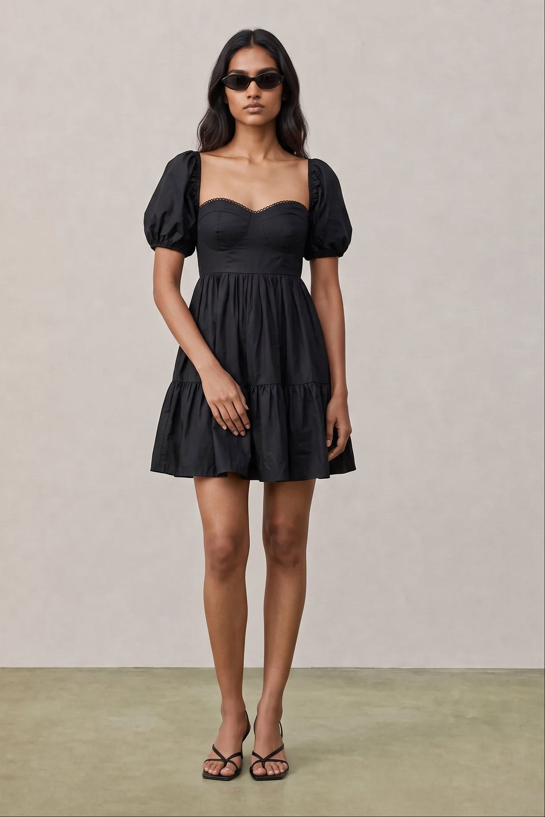 A woman modeling a black puff-sleeve mini dress with a sweetheart neckline and tiered skirt, standing against a neutral beige studio backdrop.