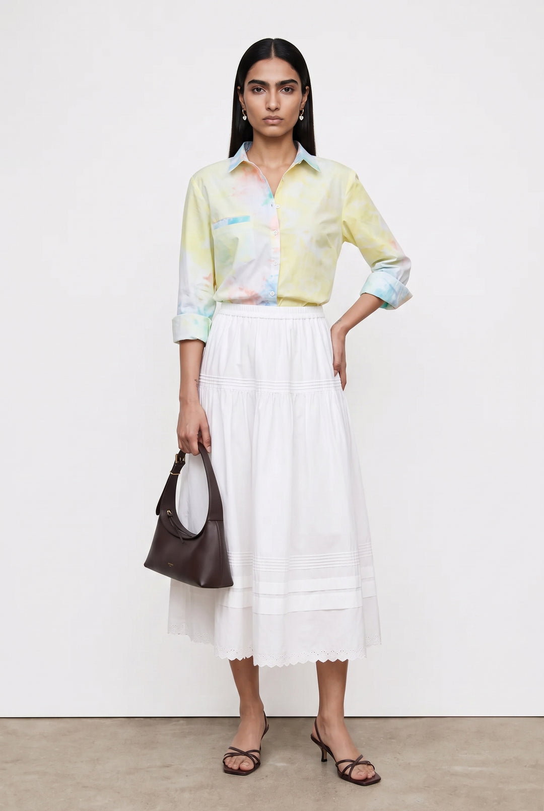 A model stands against a plain white studio wall wearing a pastel tie-dye button-up shirt, a long white pleated skirt, and black strappy heels, while holding a dark brown handbag.
