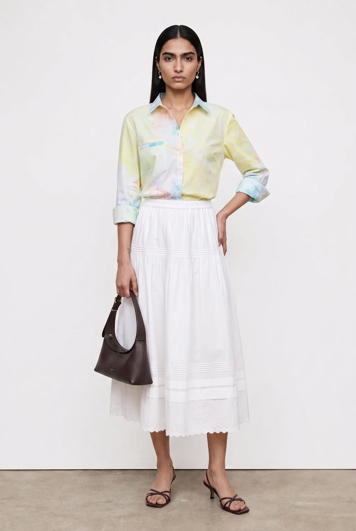 A model stands against a plain white studio wall wearing a pastel tie-dye button-up shirt, a long white pleated skirt, and black strappy heels, while holding a dark brown handbag.