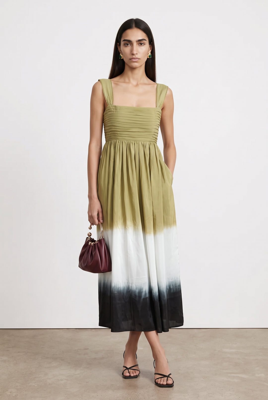 A woman models a sleeveless midi dress with a square neckline, ruched bodice, and a tie-dye ombre skirt transitioning from olive green to white and black. She is standing against a plain off-white studio wall on a light concrete floor, holding a small burgundy handbag.