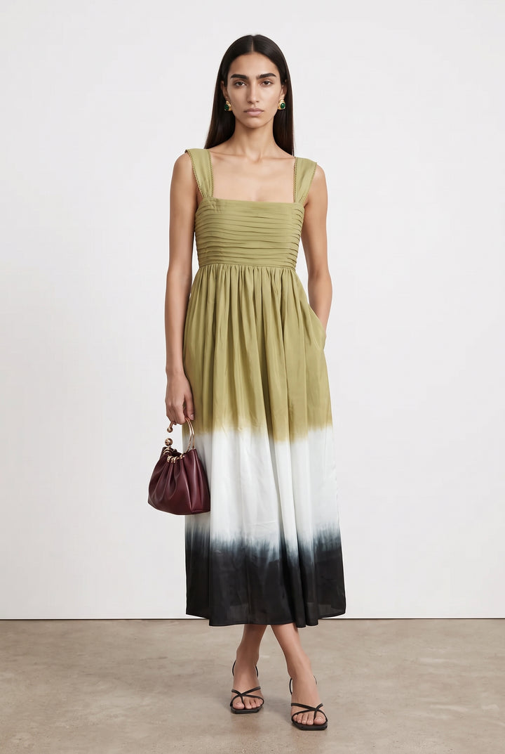 A woman models a sleeveless midi dress with a square neckline, ruched bodice, and a tie-dye ombre skirt transitioning from olive green to white and black. She is standing against a plain off-white studio wall on a light concrete floor, holding a small burgundy handbag.