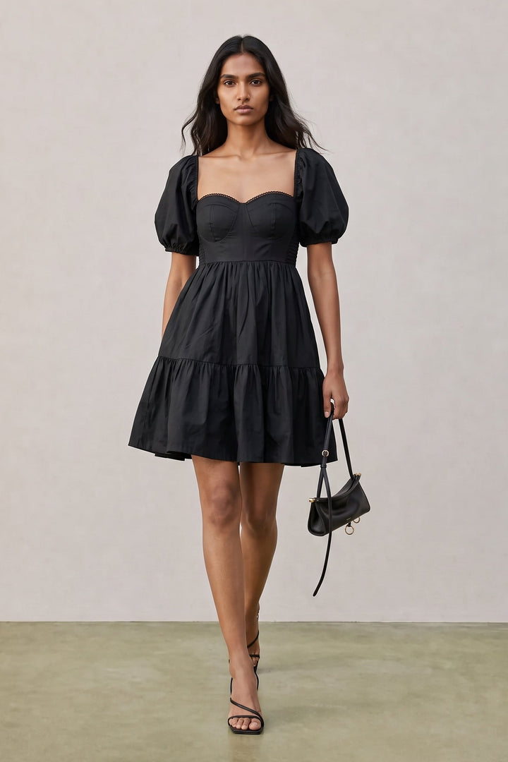 A woman models a black tiered mini dress with puff sleeves and a sweetheart neckline, holding a small black handbag against a neutral studio backdrop.