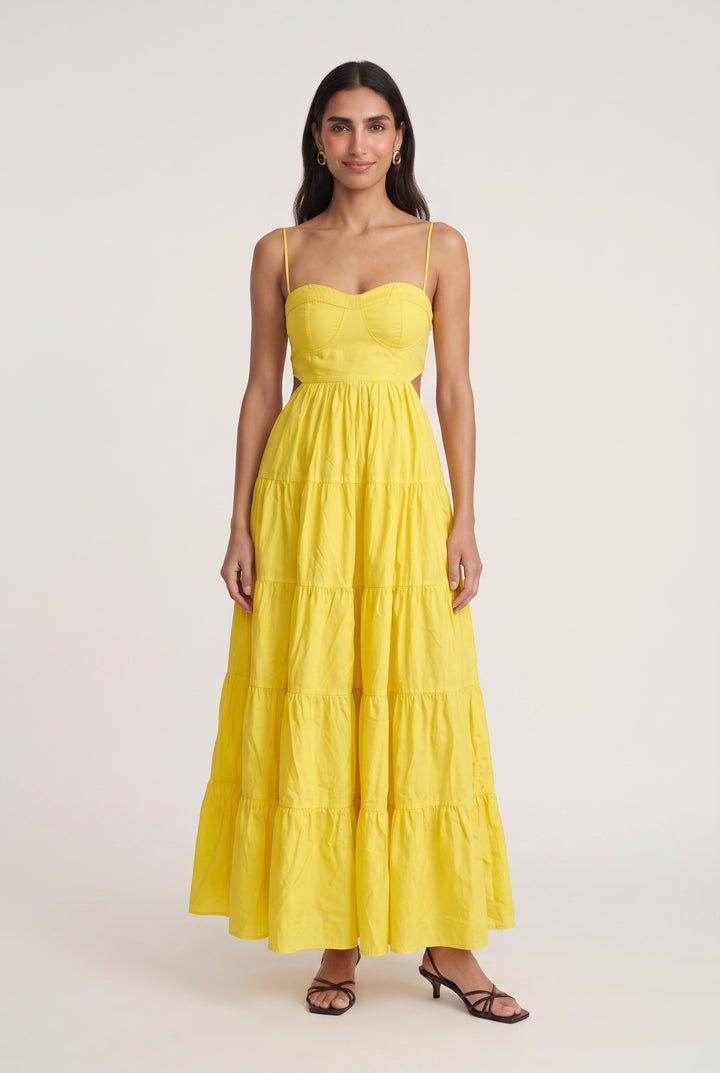 A woman modeling a bright yellow tiered maxi dress with spaghetti straps and a sweetheart neckline, standing against a plain light gray studio background.