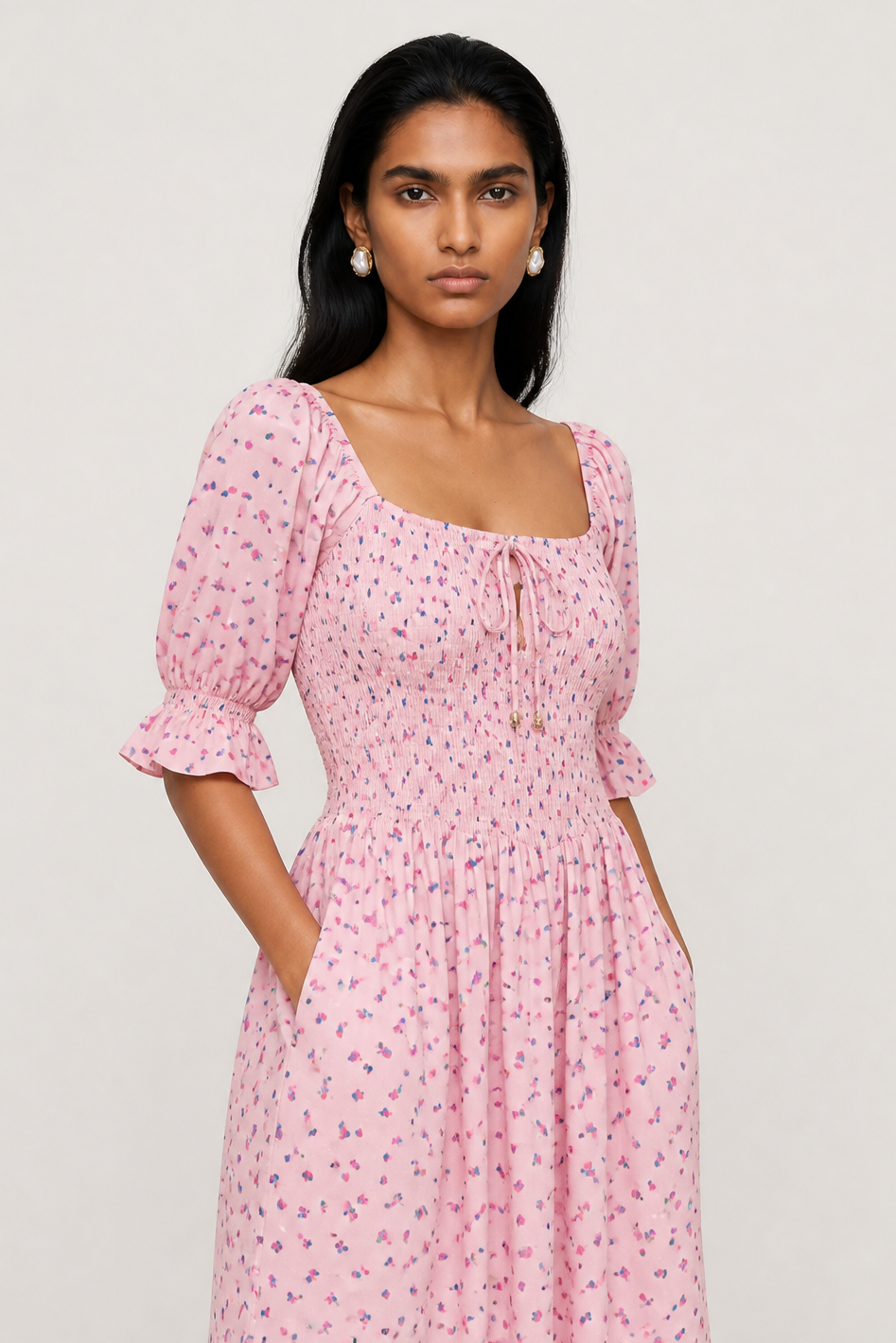A woman modeling a light pink puff-sleeve mini dress with a small blue and purple floral print, square neckline, and front tie detail, set against a plain light gray studio background.