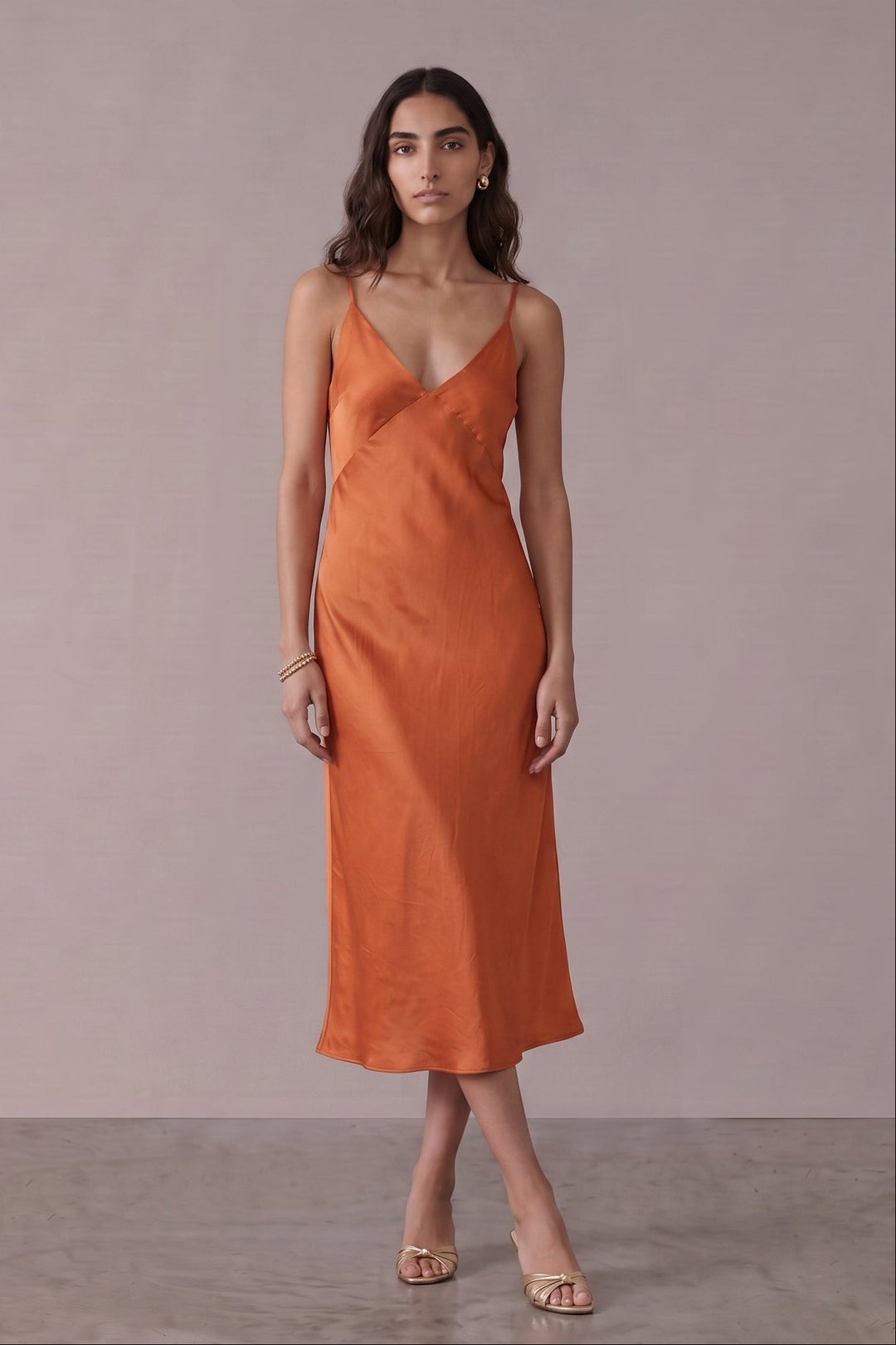A woman modeling a sleeveless, wrap-style midi dress in a warm rust orange color, standing against a neutral, textured light gray studio backdrop.