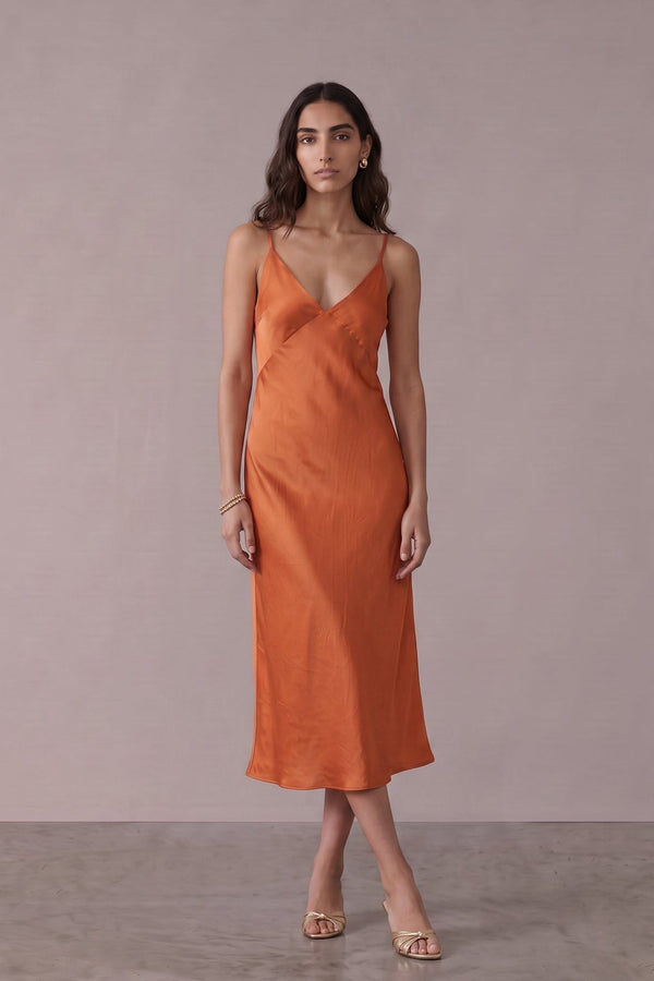A woman modeling a sleeveless, wrap-style midi dress in a warm rust orange color, standing against a neutral, textured light gray studio backdrop.