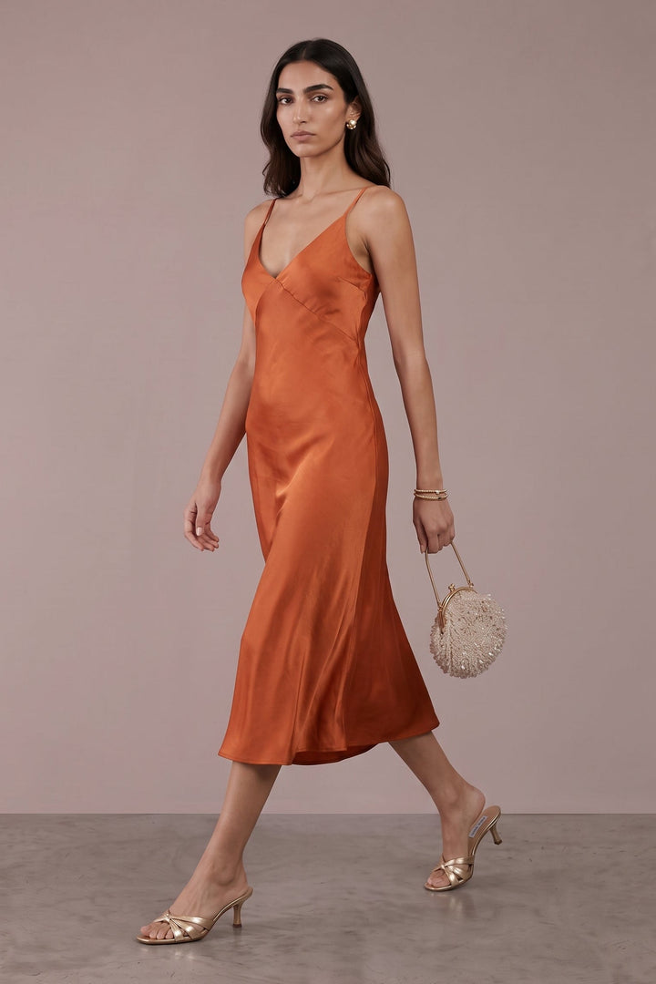 A woman models a satin-like rust-orange midi slip dress with thin straps, paired with gold heeled sandals and a small woven handbag, set against a solid mauve studio backdrop.