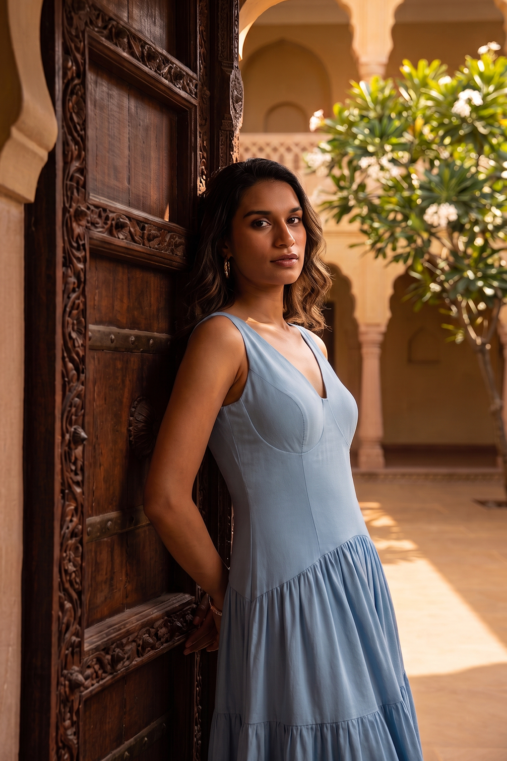 A woman wearing a light blue sleeveless tiered dress leans against a carved wooden door in a sunlit courtyard with arches and greenery.