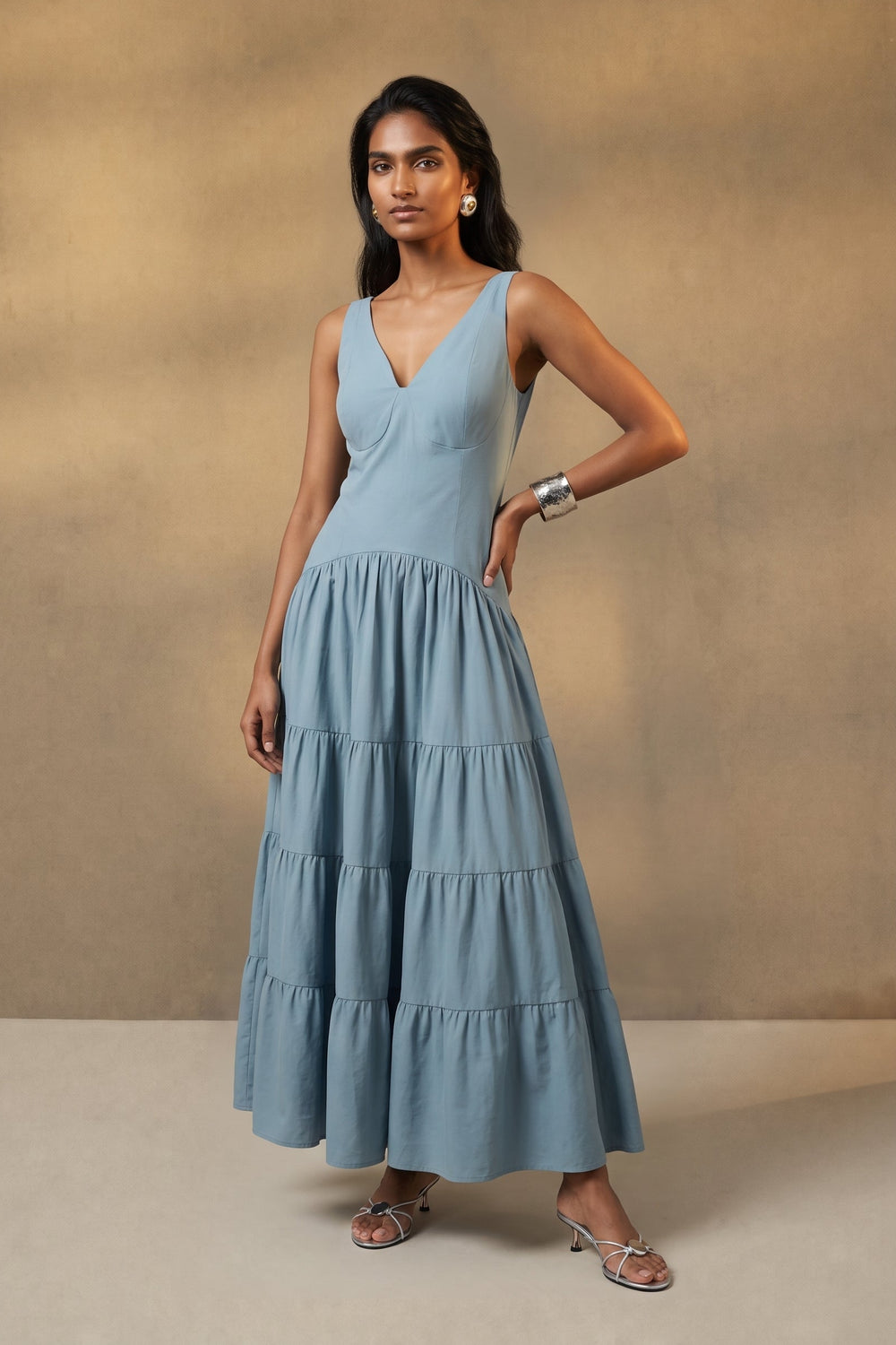 A woman modeling a light blue sleeveless tiered maxi dress against a textured beige studio backdrop.