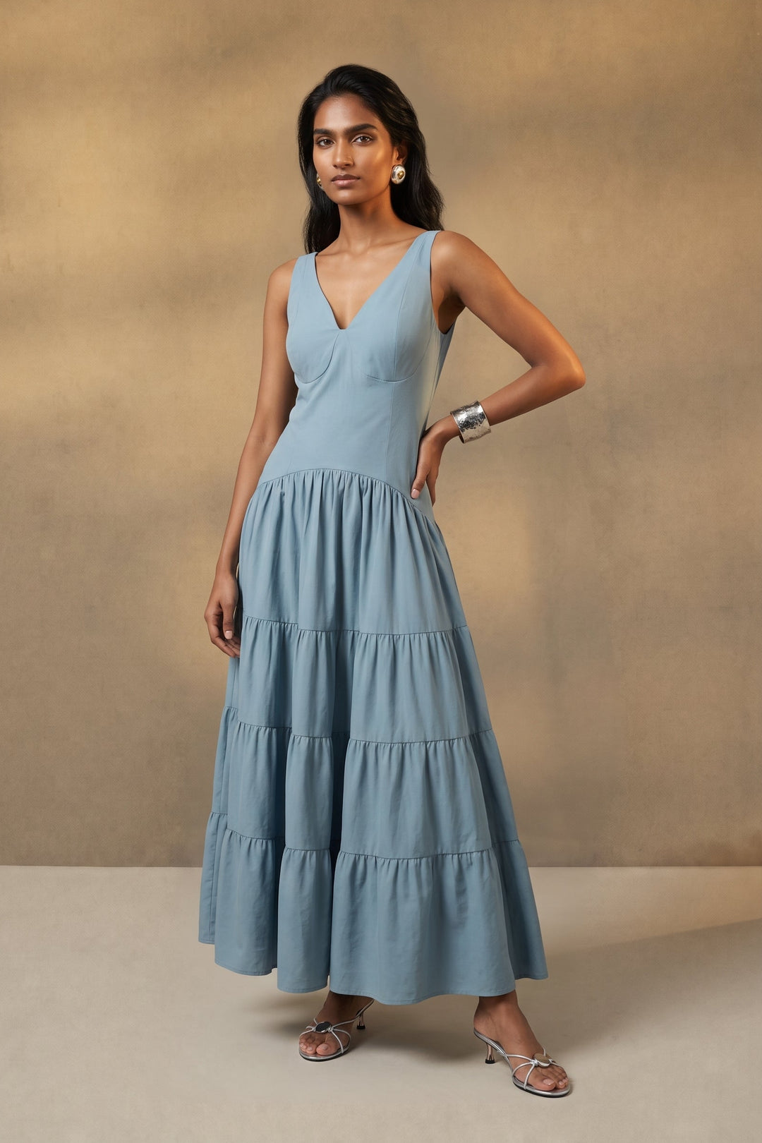A woman modeling a light blue sleeveless tiered maxi dress against a textured beige studio backdrop.