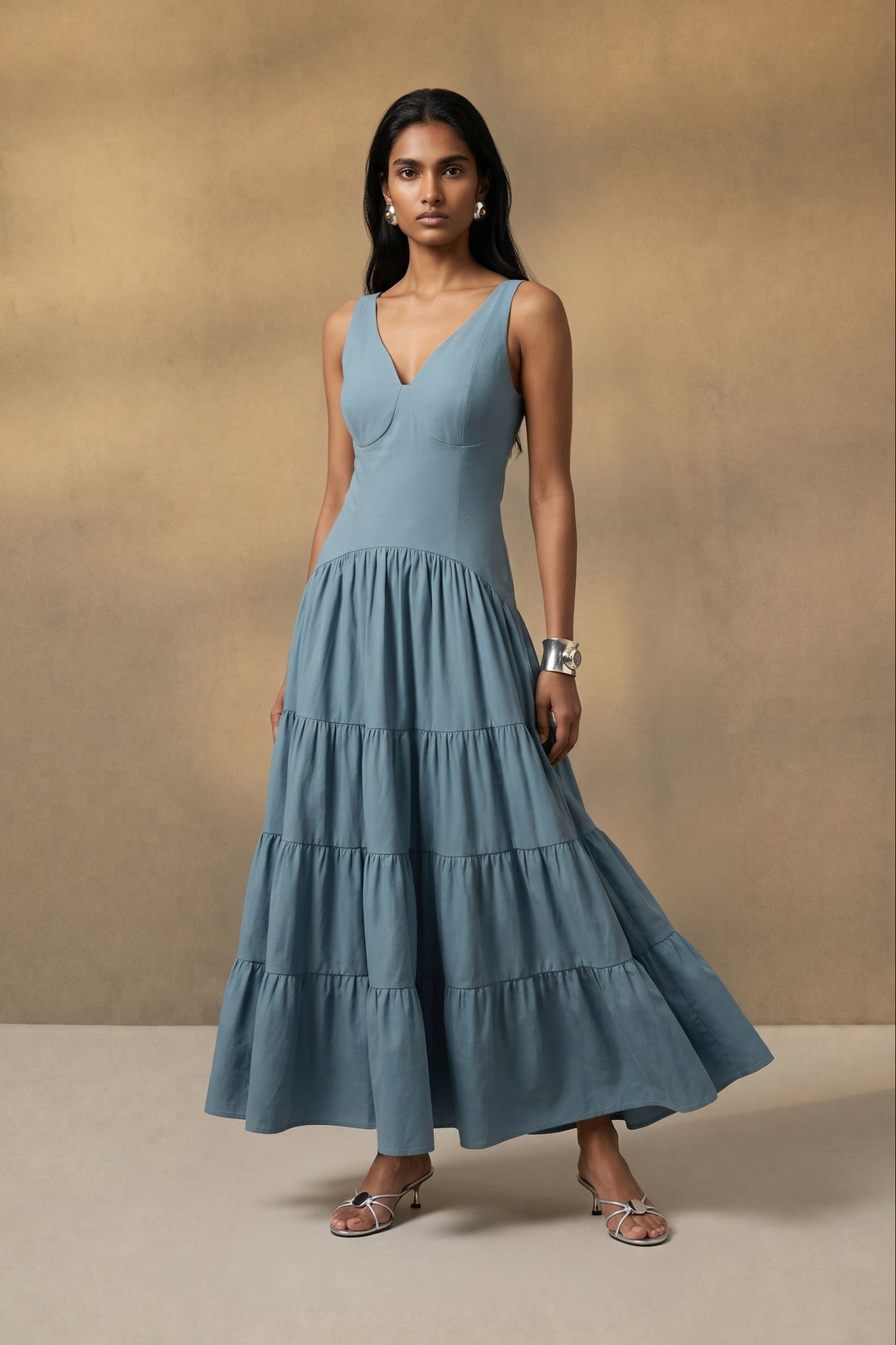A woman modeling a sleeveless, tiered maxi dress in dusty blue against a textured beige studio backdrop.