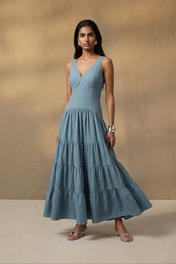 A woman modeling a sleeveless, tiered maxi dress in dusty blue against a textured beige studio backdrop.