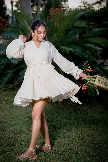 A woman in a white mini dress with long sleeves and a V-neck neckline, standing in a garden setting.