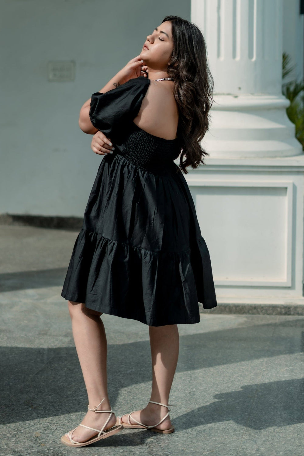 A woman wearing a black tiered off-the-shoulder midi dress and beige strappy flat sandals, posing outdoors in front of a white architectural column.