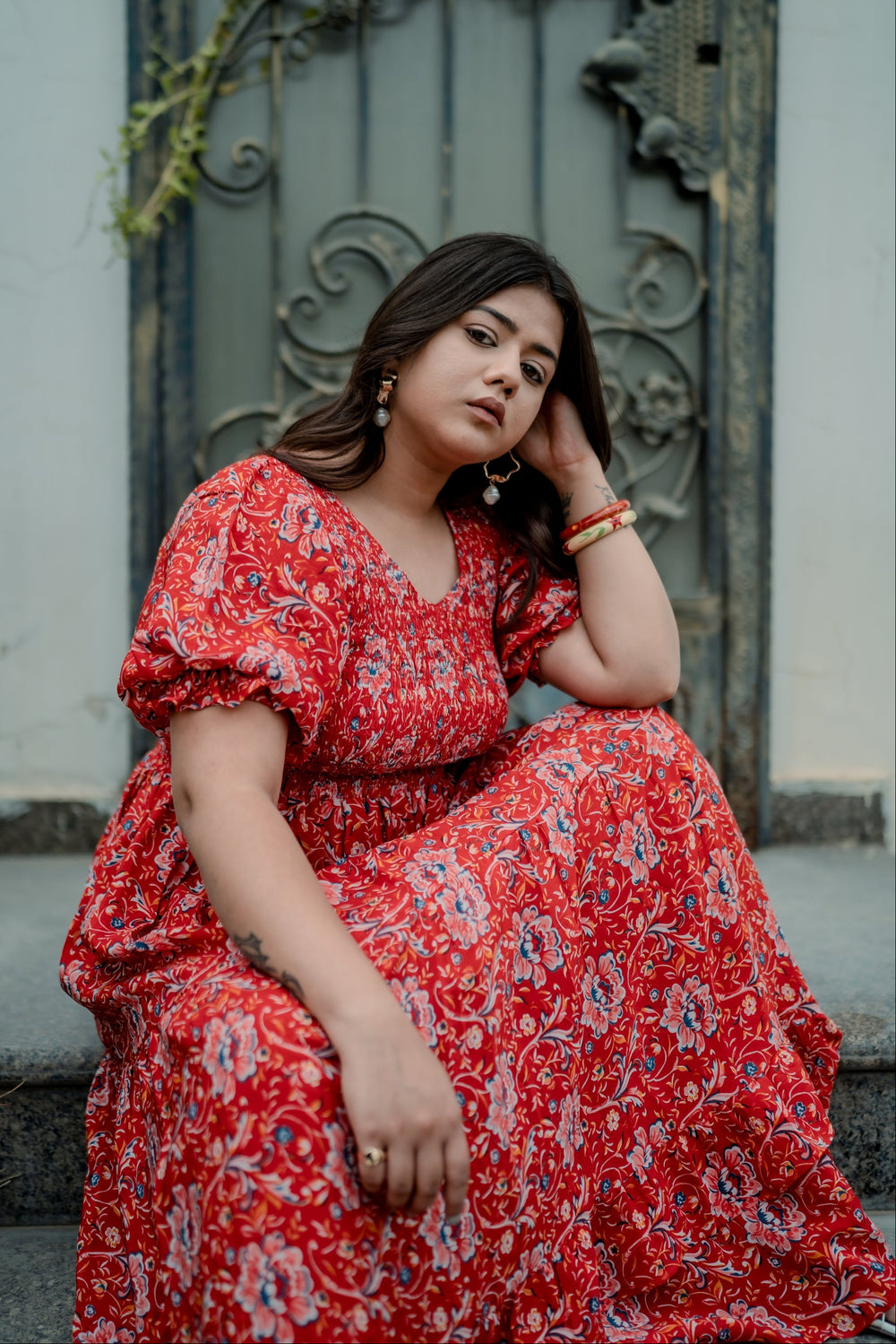 A woman sitting on a doorstep wearing a red floral printed maxi dress with short puff sleeves and a tiered skirt.