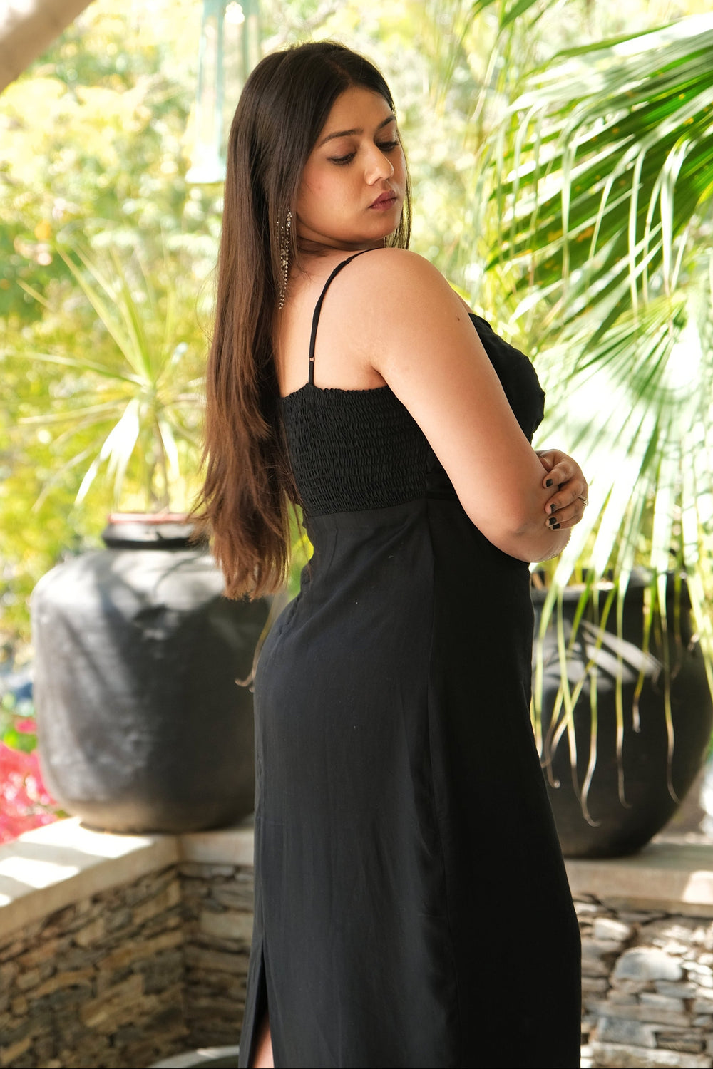 A woman wearing a black spaghetti-strap maxi dress with a textured bodice, standing in a tropical garden setting with potted plants and stone walls.