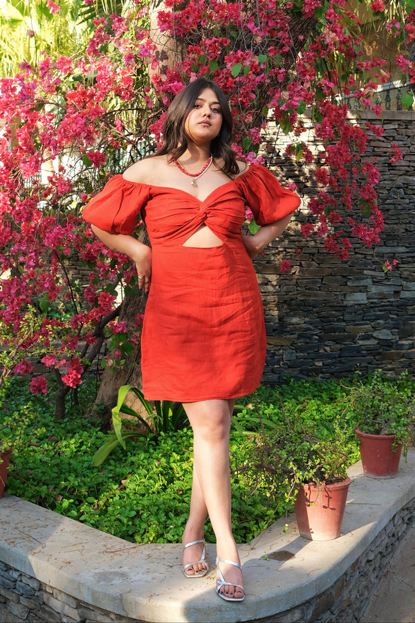 A woman wearing a bright red off-the-shoulder mini dress with a front cutout, standing outdoors in a garden setting with pink bougainvillea flowers and a stone wall behind her.