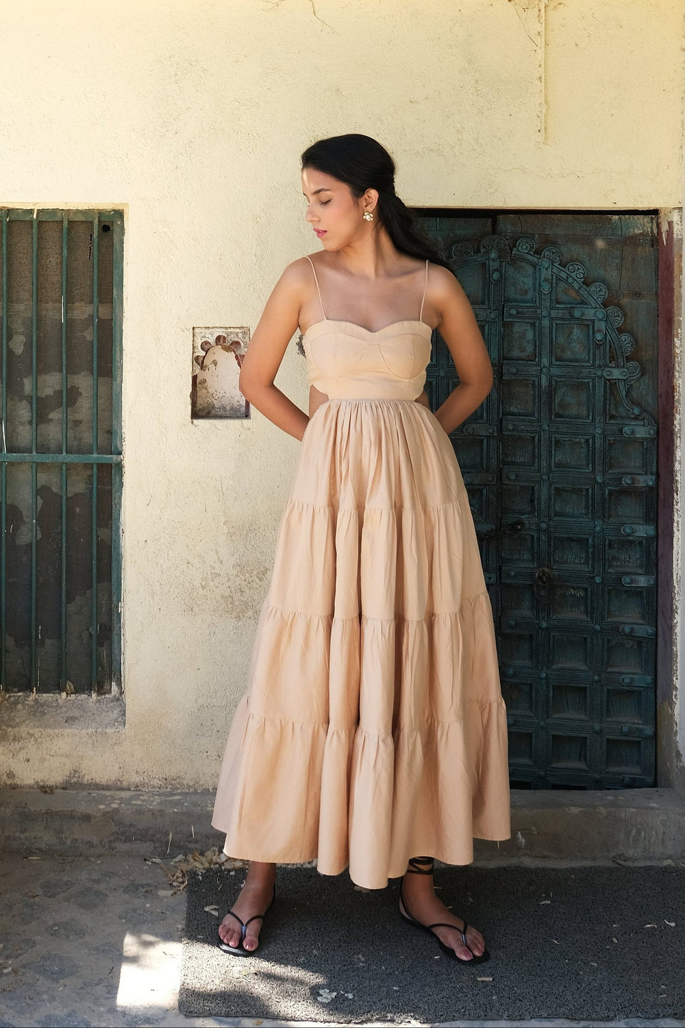 A woman wearing a strapless beige maxi dress stands in front of a rustic white stucco building with green doors and white flowers hanging over the roof.