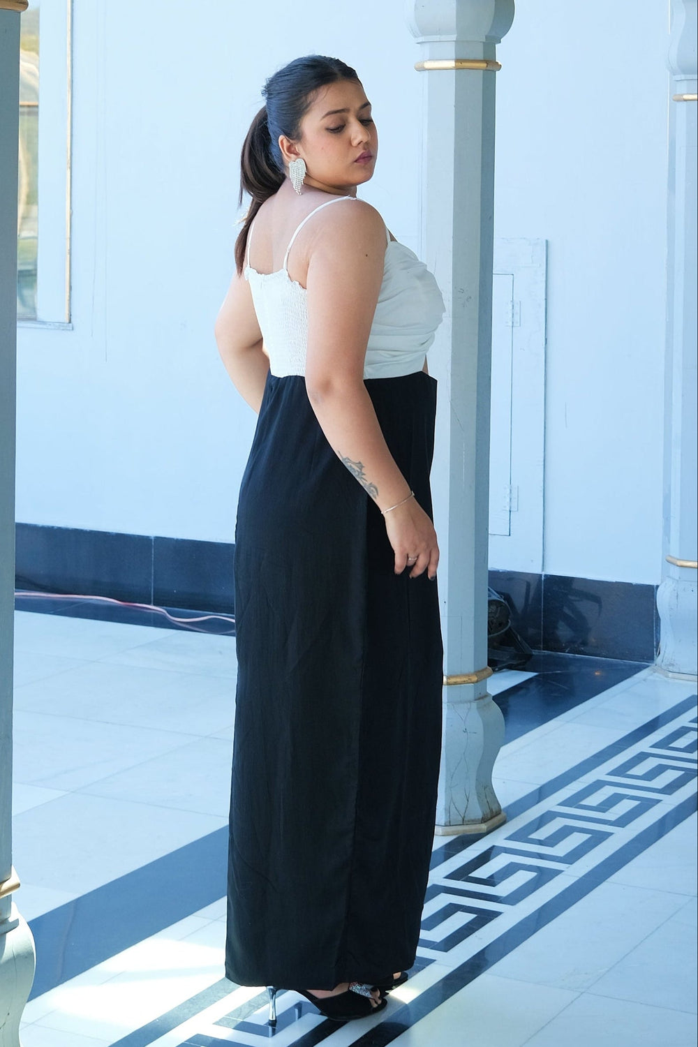 A woman stands in profile wearing a sleeveless white top and a long black skirt inside a decorative architectural corridor with arched openings and pale columns.