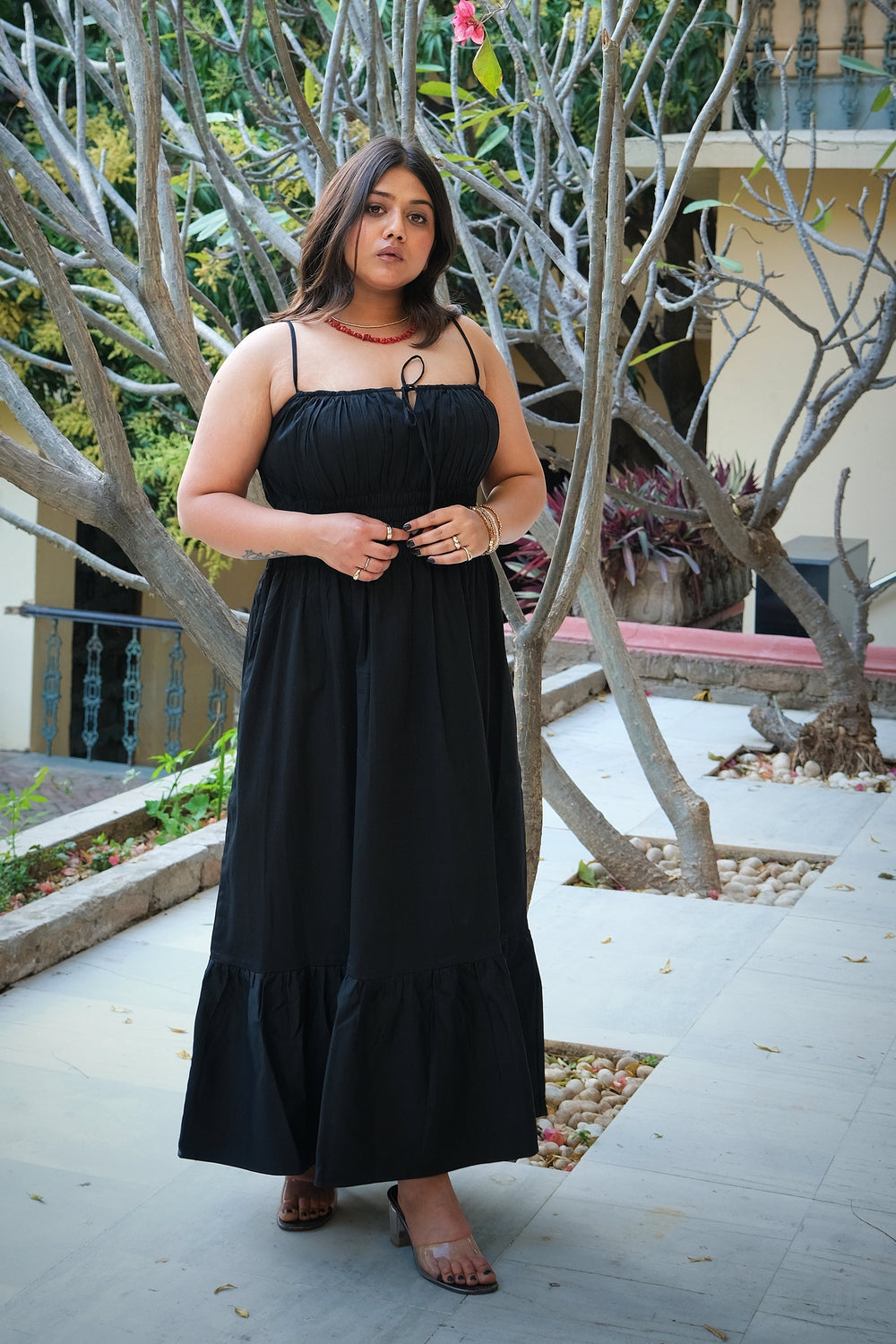A woman wearing a black spaghetti-strap maxi dress with a tie-neck detail, standing outdoors on a paved walkway surrounded by trees.