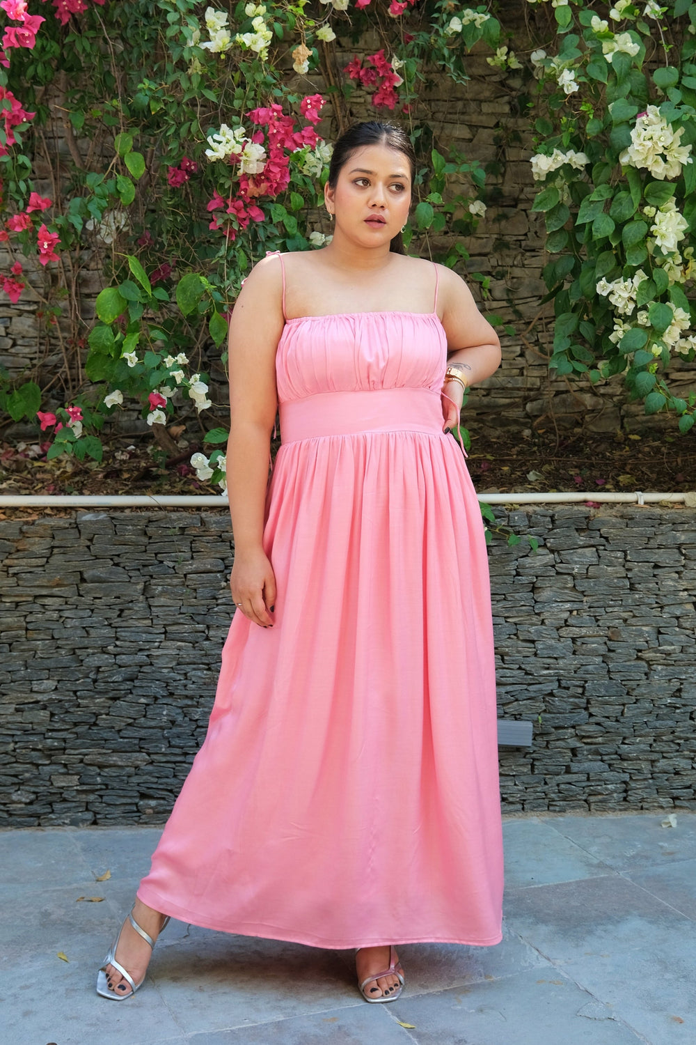 A woman wearing a strapless pink maxi dress stands outdoors in front of a stone wall covered with green vines and pink and white flowers.