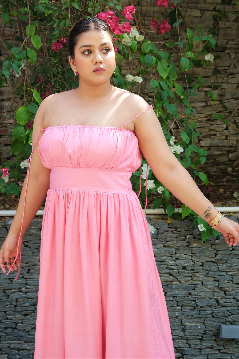 A woman wearing a light pink strapless maxi dress stands outdoors in front of a stone wall covered with green vines and pink flowers.