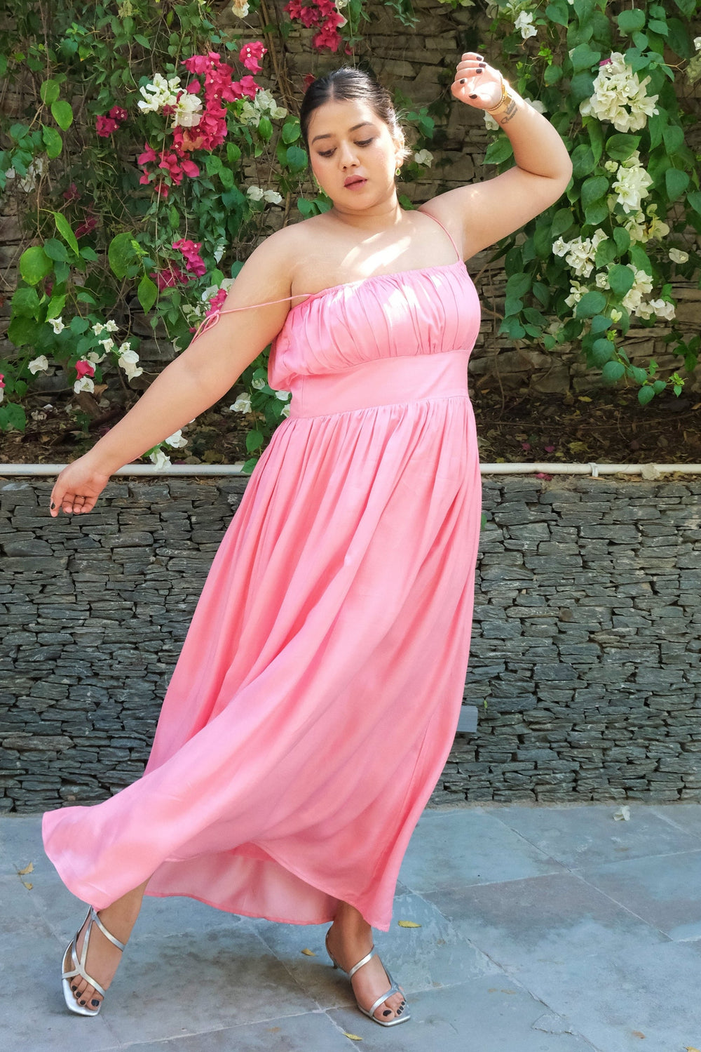 A woman wearing a strapless pink maxi dress poses outdoors in front of a stone wall covered in green vines and pink and white flowers.