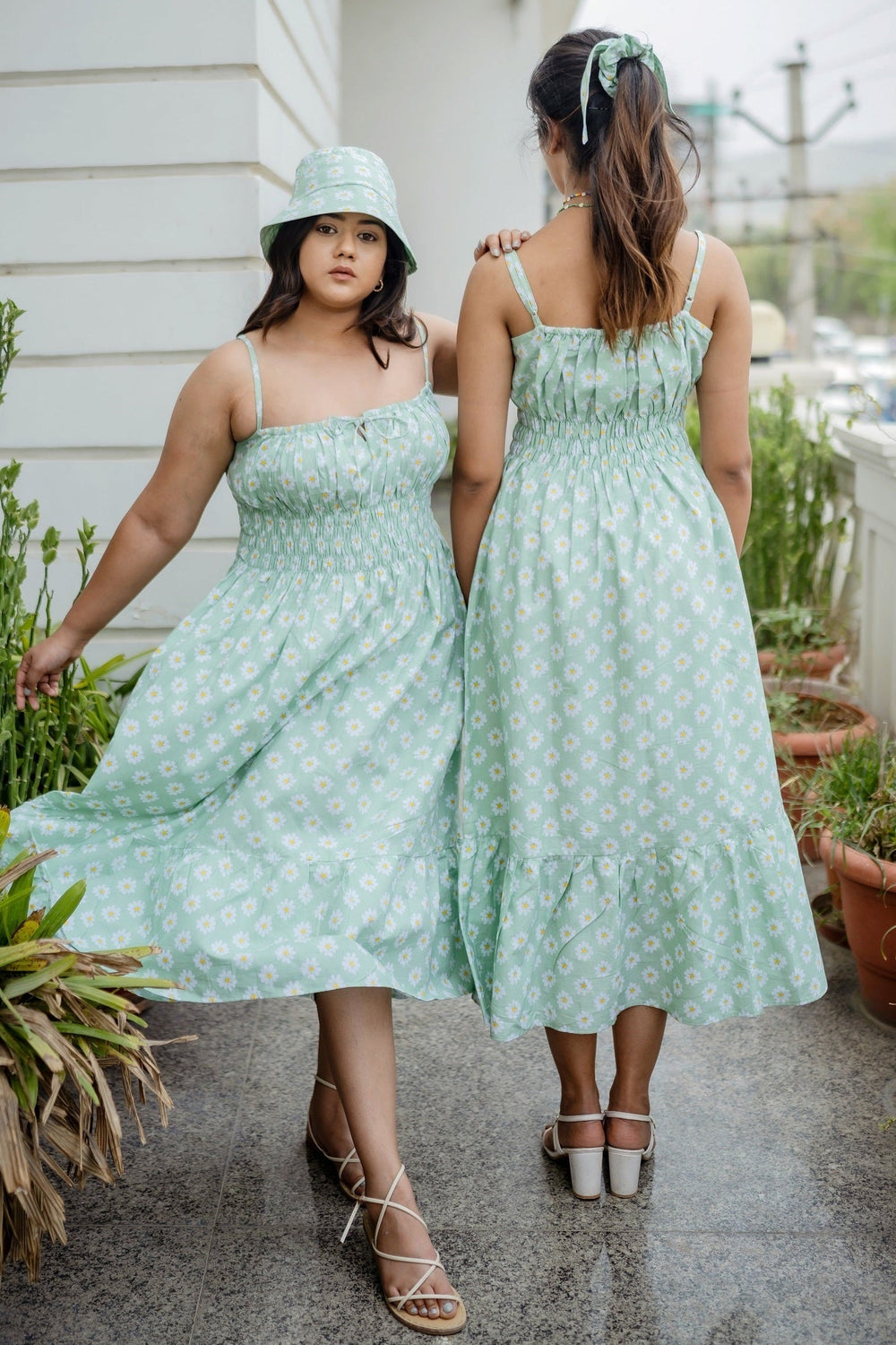 Two women modeling matching mint green midi dresses with a white floral pattern, standing outdoors on a patio. One woman is shown from the front wearing a matching bucket hat, and the other is shown from the back.