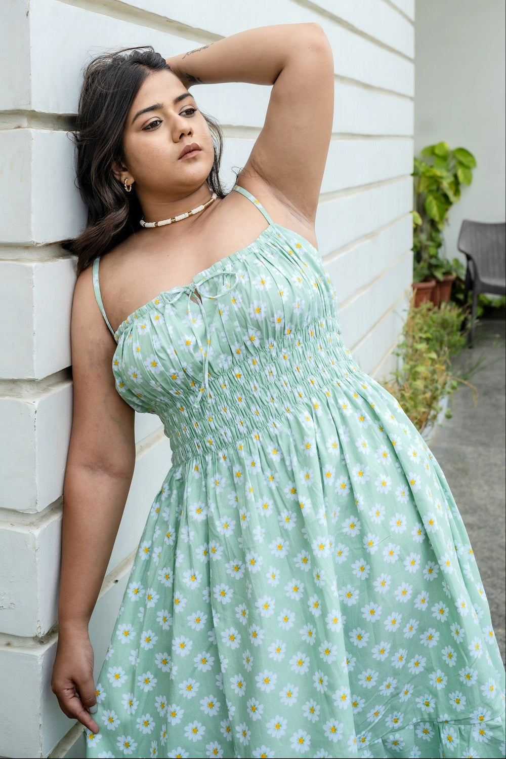 A woman wearing a light mint green spaghetti-strap maxi dress with a white and yellow daisy floral print, leaning against a white brick wall.